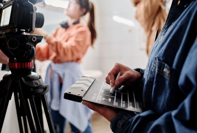 Close-up of hands holding clapperboard while filming with professional camera in indoor setting. Background showing blurred figure working with equipment